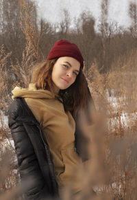 Stylish young woman in burgundy hat and gorich-colored hoodie stands in dry tall grass reeds