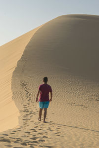 Rear view of man walking on sand dune at desert against clear sky