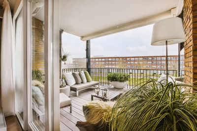 Comfortable sofas with cushions and armchairs placed near wooden table on terrace with glass entrance door of modern residential building