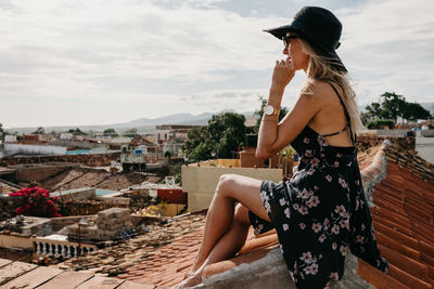 Side view of young woman looking at cityscape while sitting on rooftop against sky during sunny day