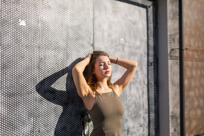 Beautiful young woman with hand in hair standing against wall