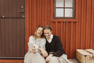 Female friends laughing while taking selfie on mobile phone against cottage