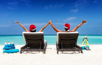 People in santa hat sitting on lounge chair at beach against sky