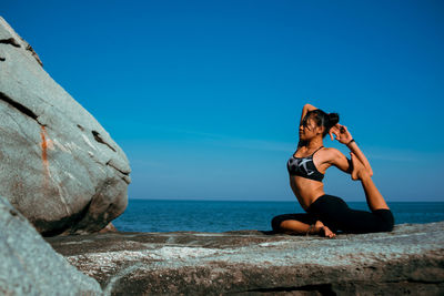 Young woman practicing yoga on rock at beach against blue sky