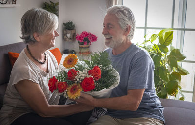 Smiling senior couple with bouquet embracing while sitting on sofa at home