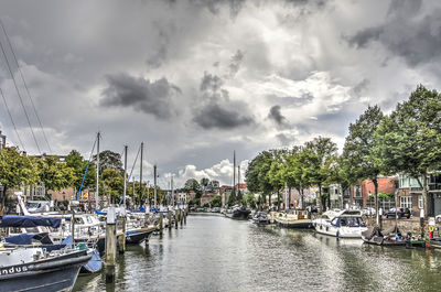 Sailboats moored on canal in city against cloudy sky