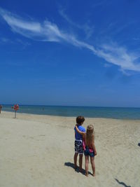 Rear view of siblings standing on beach against blue sky