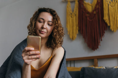 Young woman using phone, relaxing in bed, checking social media after waking up