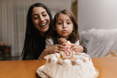 Girl blowing candle over birthday cake while sitting with mother at home