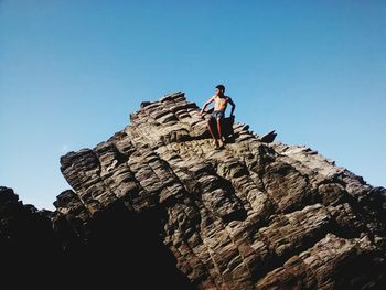 Low angle view of shirtless boy sitting on rock formation against clear blue sky