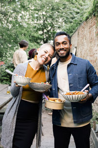 Portrait of smiling male and female friends holding food bowls while standing in back yard