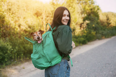 A smiling girl is holding a green backpack on her shoulder, from which a cute dog looks out. 