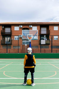 Back view anonymous sportsman in activewear standing on public sports ground with yellow ball and basketball hoop during game on street