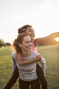 Woman giving piggyback to daughter at park against sky during sunset