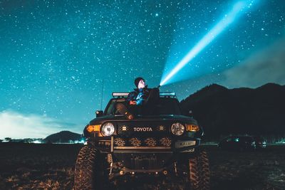 Man sitting on illuminated car against sky at night