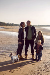 Family in a leather jacket stands along the beach with their dog in autumn