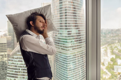 Thoughtful businessman with beard stands next to window in office against backdrop skyscrapers