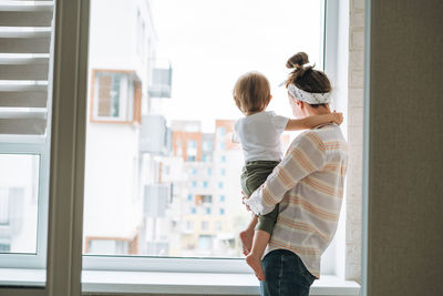 Young woman mother with baby girl daughter on window sill looking at window at home