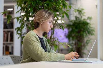 Pensive university teacher working on laptop in cozy empty library filled with green houseplants