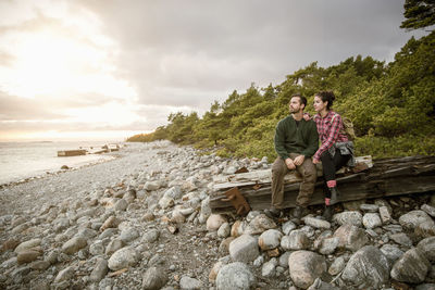 Couple sitting on wood at beach against sky