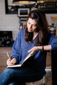 Concentrated female in casual wear resting in cozy coffee shop and writing thoughts in diary while enjoying weekend