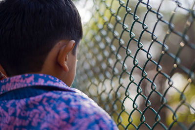 Rear view of boy looking through chainlink fence