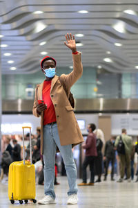 Full length of man wearing mask standing with hand raised at airport