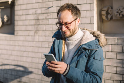 Attractive smiling male with beard, glasses and in blue jacket booking airplane tickets on website.