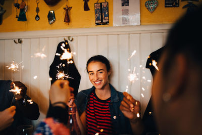 Smiling young multi-ethnic friends holding burning sparklers while enjoying in restaurant during dinner party