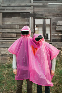 Friends wearing pink raincoat and virtual reality stimulators while standing against abandoned house