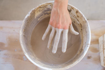 From above crop anonymous craftswoman standing near table and pulling dirty hand from bucket filled with clay in studio in daylight