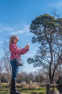 Full side view of an adult woman with sunglasses taking a picture in a park.