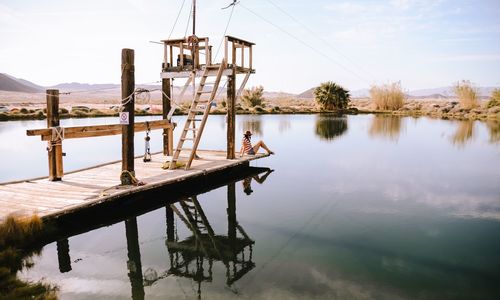 Full length of woman sitting on jetty over lake against sky
