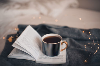 Cup of coffee on knit textile sweater in bed with paper open book with christmas glow light closeup