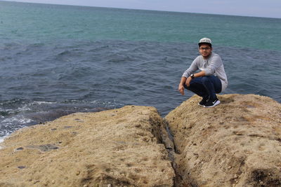 Full length portrait of man crouching on rock at beach