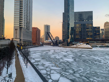 Frozen river by buildings against sky during sunset in city