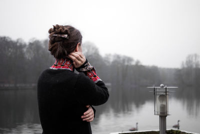 A young woman stands pensively on the terrace on a cold day and looks out over the grey lake