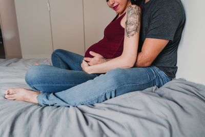 Midsection of pregnant woman sitting with male partner on bed at home