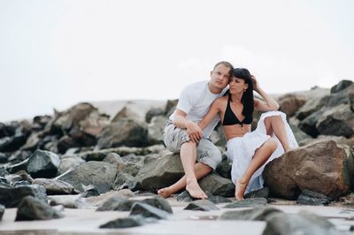 Young couple sitting on rock against sky