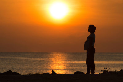 Side view of silhouette pregnant woman standing at beach during sunset