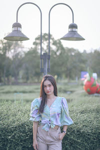 Portrait of woman standing by street lights against plants in park