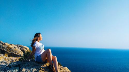 Woman sitting on rock by sea against clear blue sky