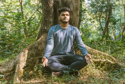 Young man meditating while sitting against tree trunk in forest