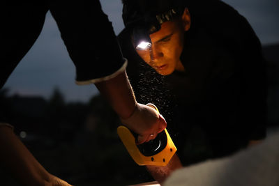 Man using saw to cut wood, yellow light from lantern, night setting, sawdust in production. 