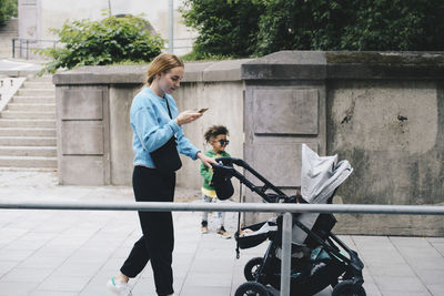 Mother with son using mobile phone while pushing baby stroller on sidewalk at city