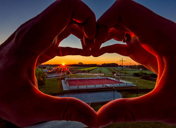 Close-up of man holding heart shape against sky during sunset