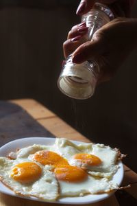 Cropped hands sprinkling pepper on fried eggs in plate