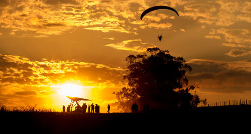 Silhouette people paragliding and hang-gliding on field against dramatic sky during sunset