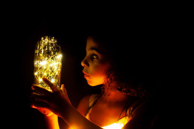 Portrait of young woman holding illuminated lighting equipment against black background