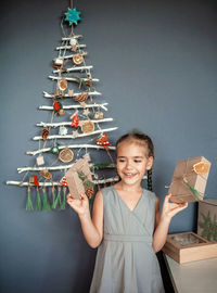Smiling girl holding gift against christmas tree hanging on wall